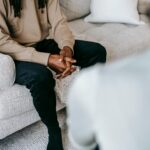 Unrecognizable ethnic male client with hands clasped sitting on sofa near blurred female therapist during psychotherapy appointment in cozy office